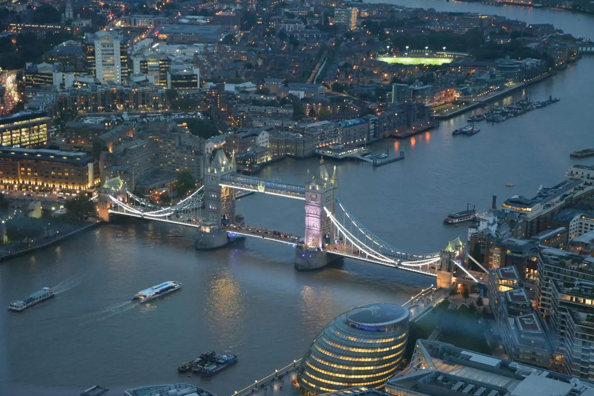 Aerial night view of Tower Bridge in London, illuminated and spanning the River Thames, with city lights in the background and boats on the water.