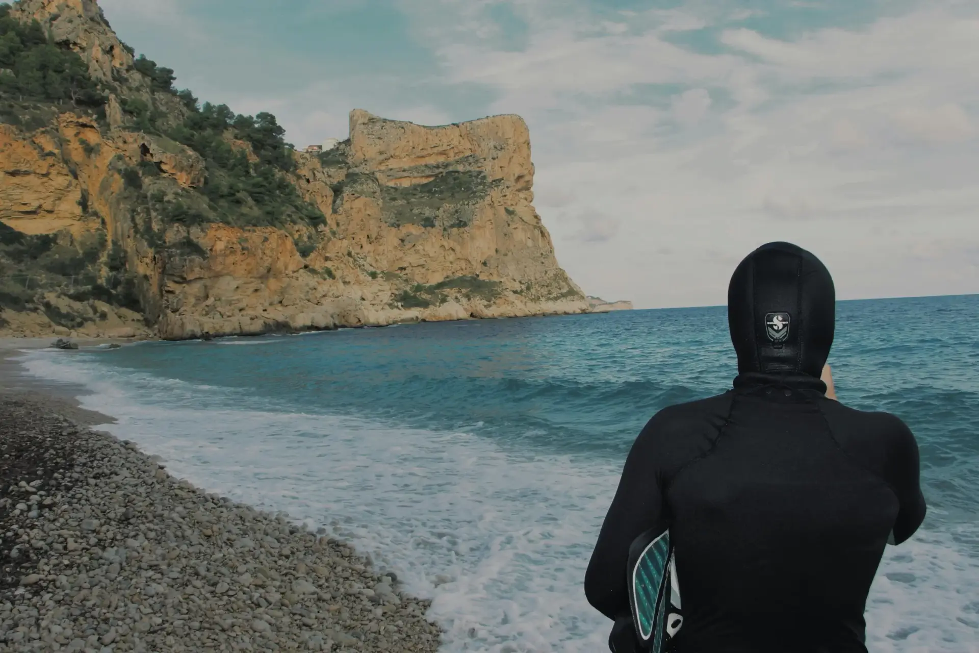 Person in a full black wetsuit looking out at a secluded Mediterranean cove with a pebble beach and clear turquoise water, ready for diving or snorkeling.