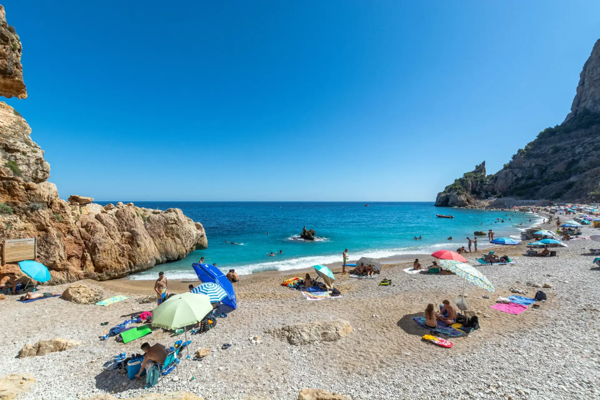 Wide view of a crowded pebble beach in a Mediterranean cove on a sunny day, with many colorful umbrellas, people enjoying the sea, and rocky cliffs.