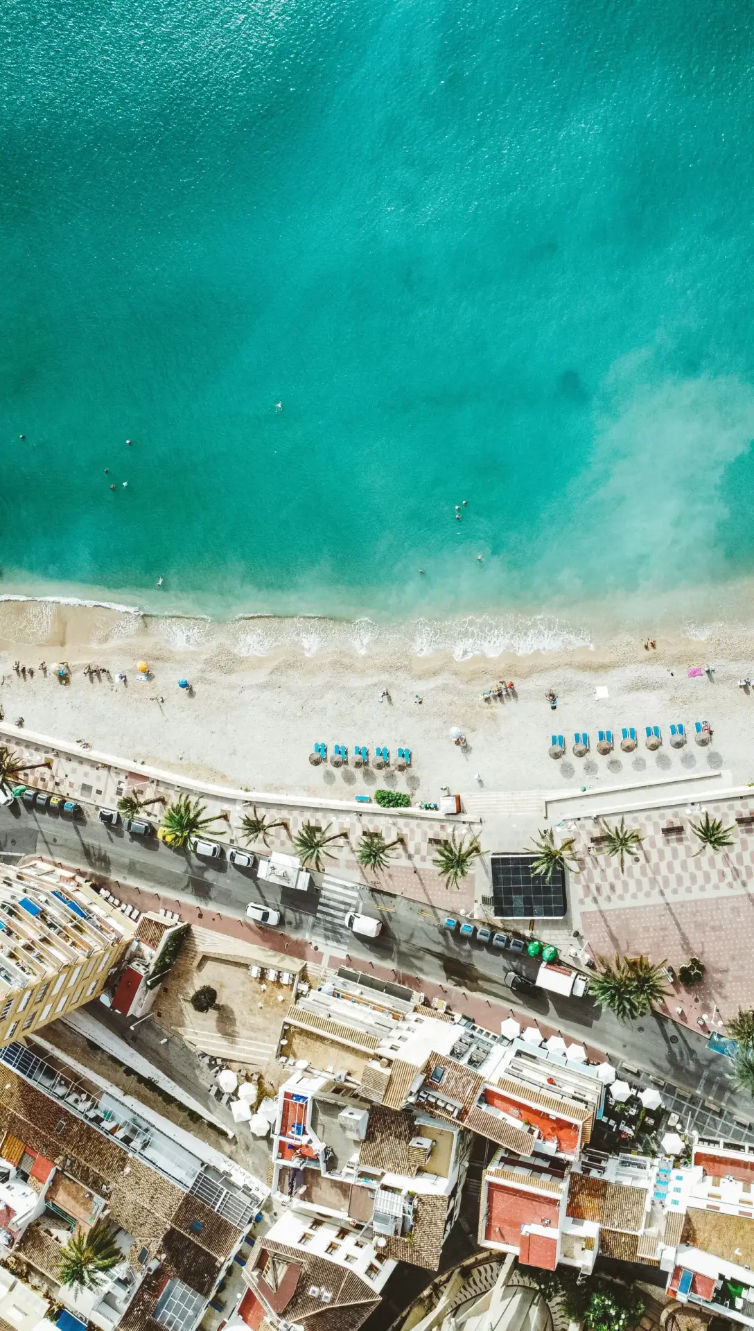 Top-down aerial view of a vibrant coastal town meeting a beach with crystal-clear turquoise water, showing the sandy bottom near the shore.
