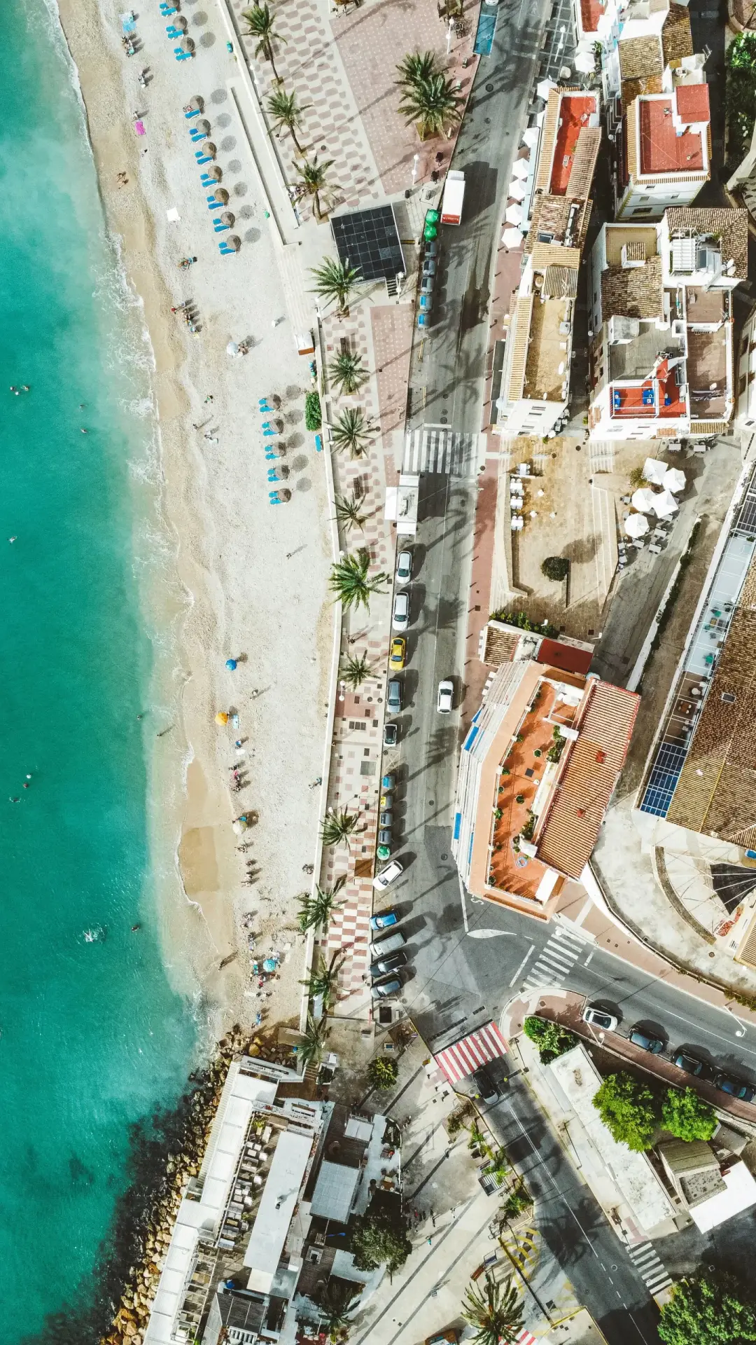 Families enjoying the sun and turquoise waters at Arenal Beach in Javea, an aerial view showing the clear sea and sandy shore.
