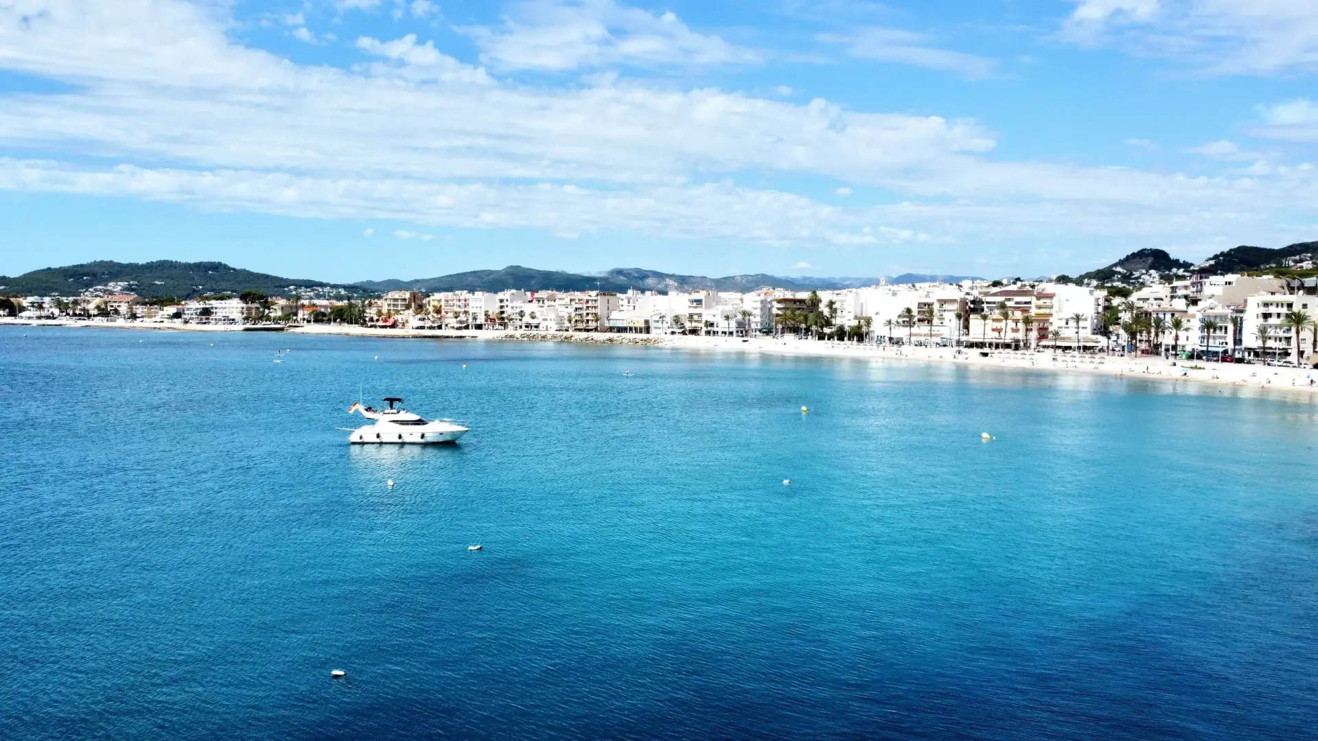 Scenic view of a Mediterranean bay with a sandy beach, clear turquoise waters, a white boat anchored, and a coastal town lining the shore.