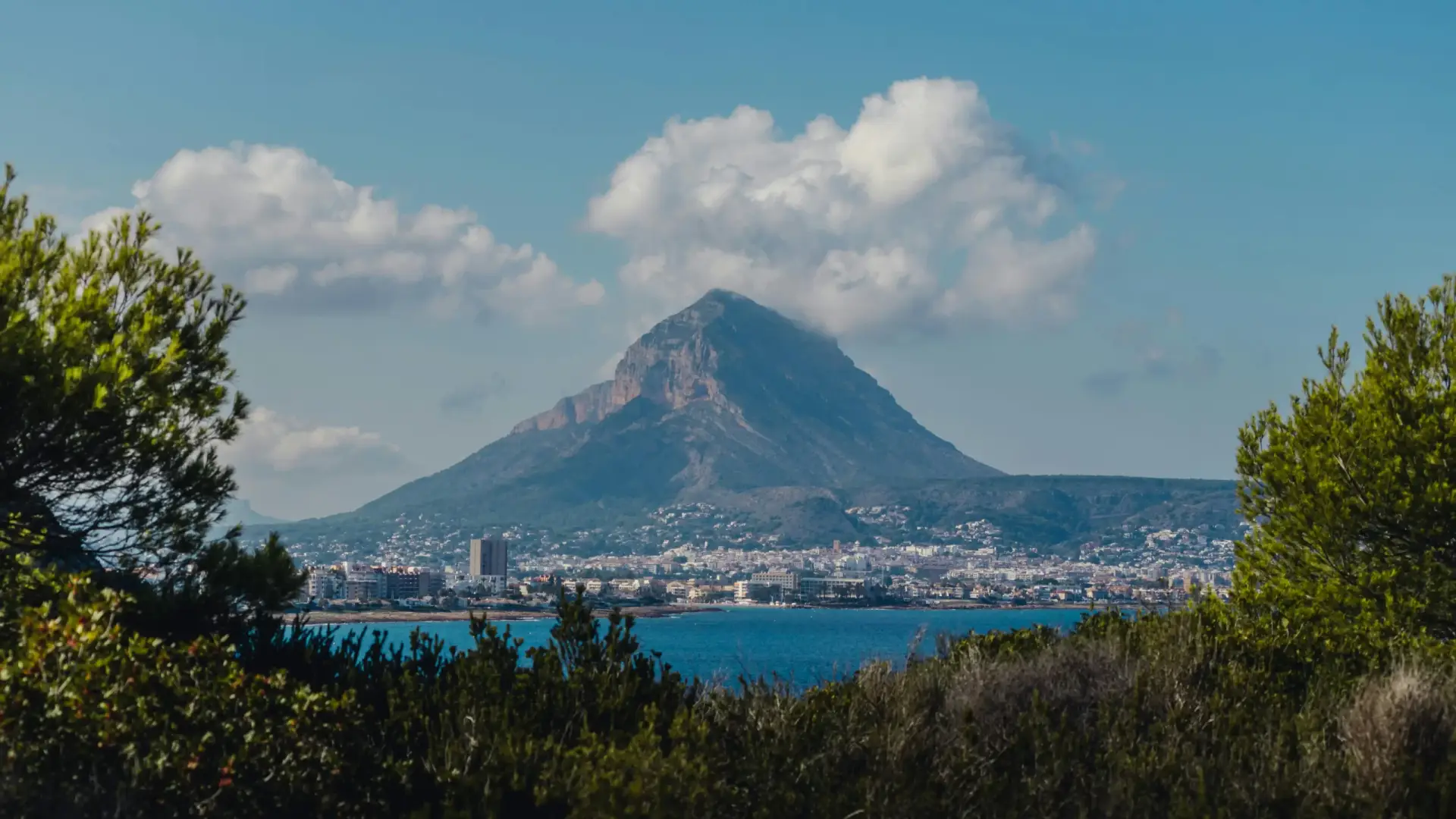Scenic aerial view of Javea's coastline featuring the Montgó Mountain, the town, and the Mediterranean Sea, framed by foreground trees.