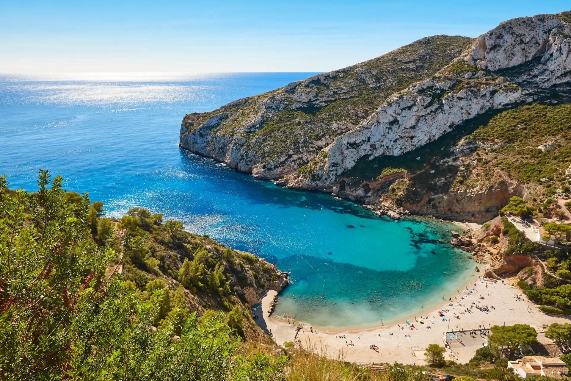 Aerial view of the stunning Cala Granadella beach in Javea, Spain, showcasing its turquoise waters, pebble beach, and surrounding pine-covered cliffs.