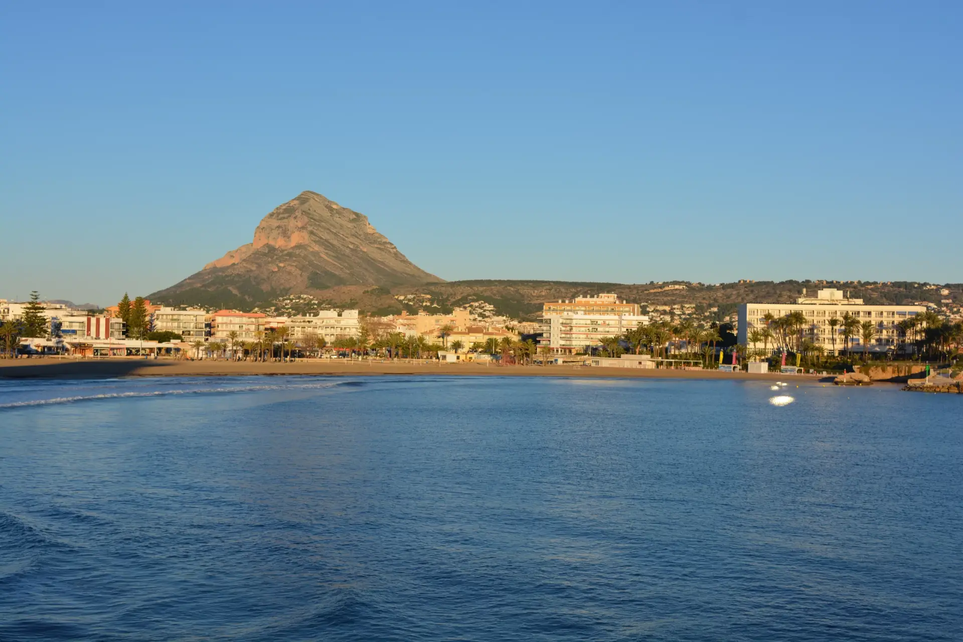 Panoramic waterfront view of Arenal Beach in Javea, Costa Blanca, showing the sandy beach, town, Montgó mountain, and boats on the sea.