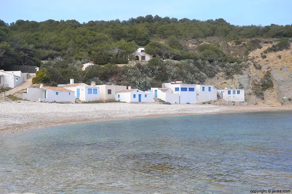 Traditional white fisherman's houses with blue doors and windows lining the pebble beach of Cala de la Barraca (Portitxol) in Javea, Spain, with the Mediterranean sea in the foreground.