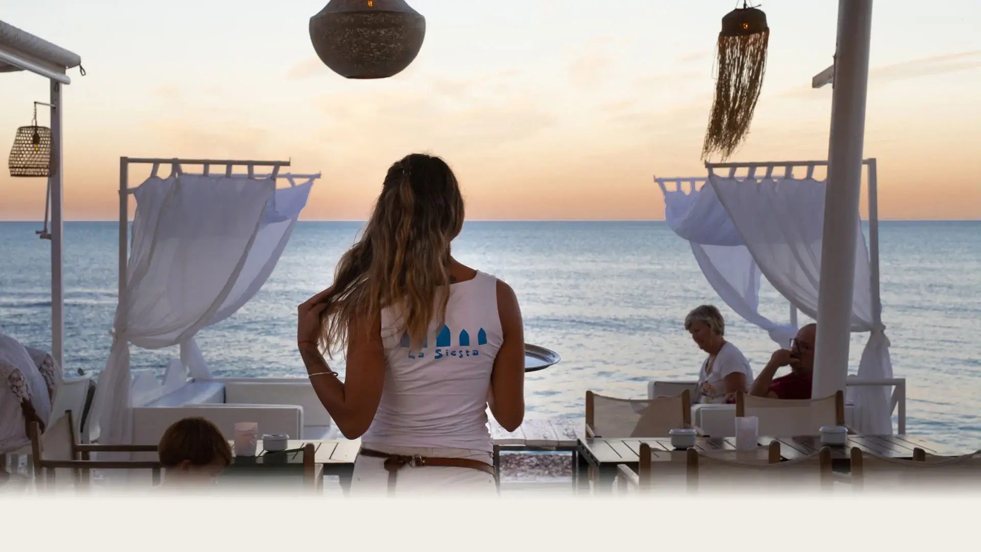 Woman enjoying the evening ambiance at La Siesta beach club in Javea, with soft lighting, draped cabanas, and outdoor seating.