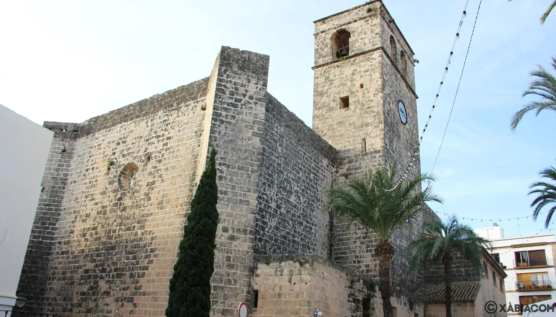 The historic San Bartolomé Church in Javea, Spain, showcasing its Gothic stone facade, bell tower, and palm trees in the foreground.