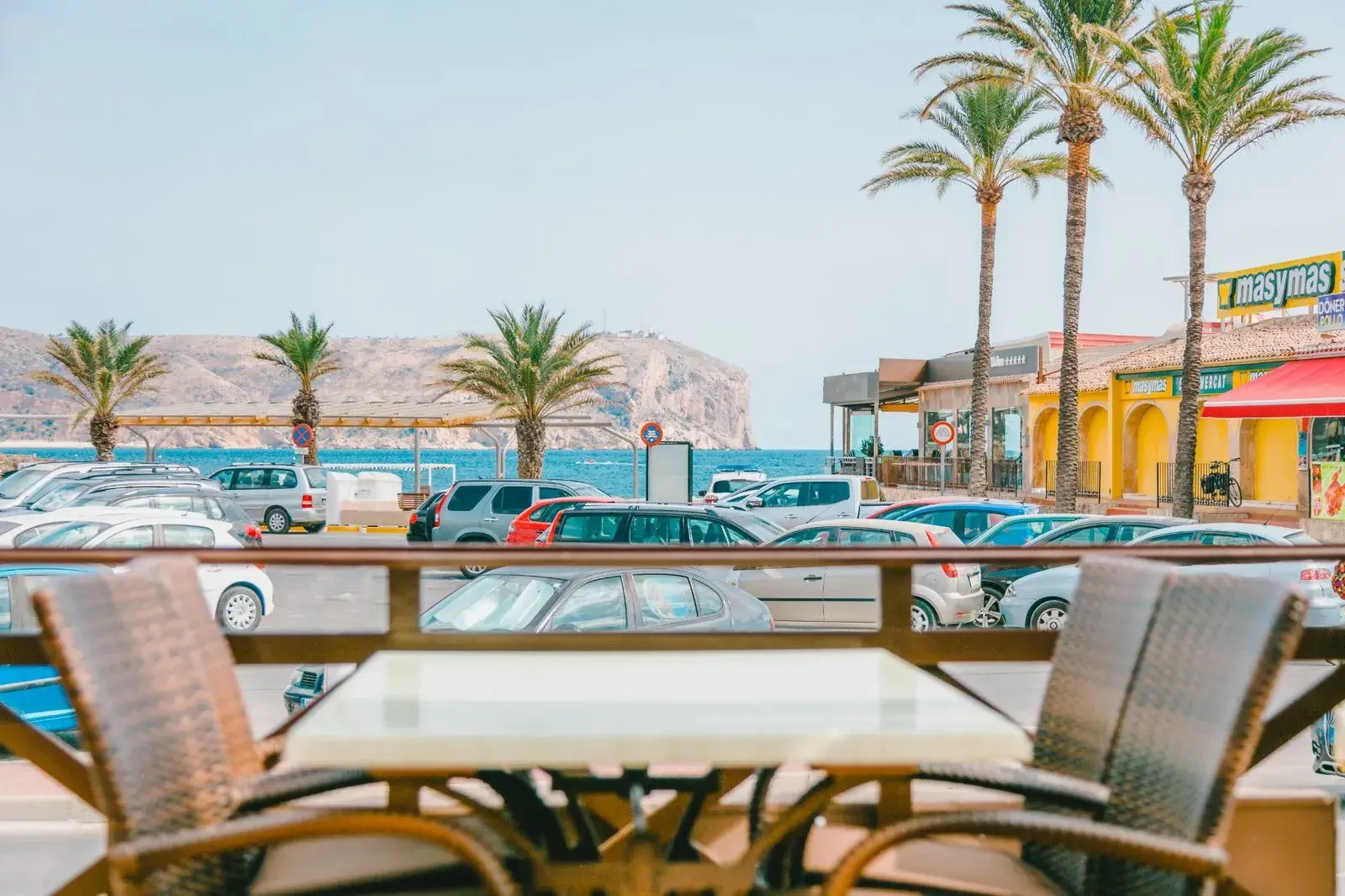 Street view from an outdoor seating area showing parking, shops, palm trees, and coastline