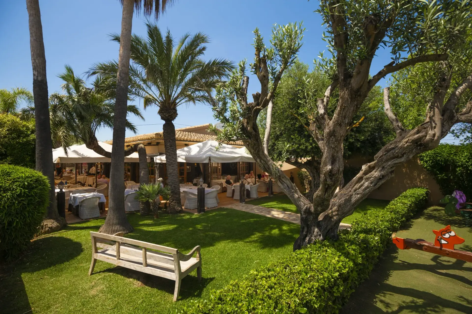 Sunny garden and patio seating area of a restaurant, possibly Restaurante Masena, with mature trees
